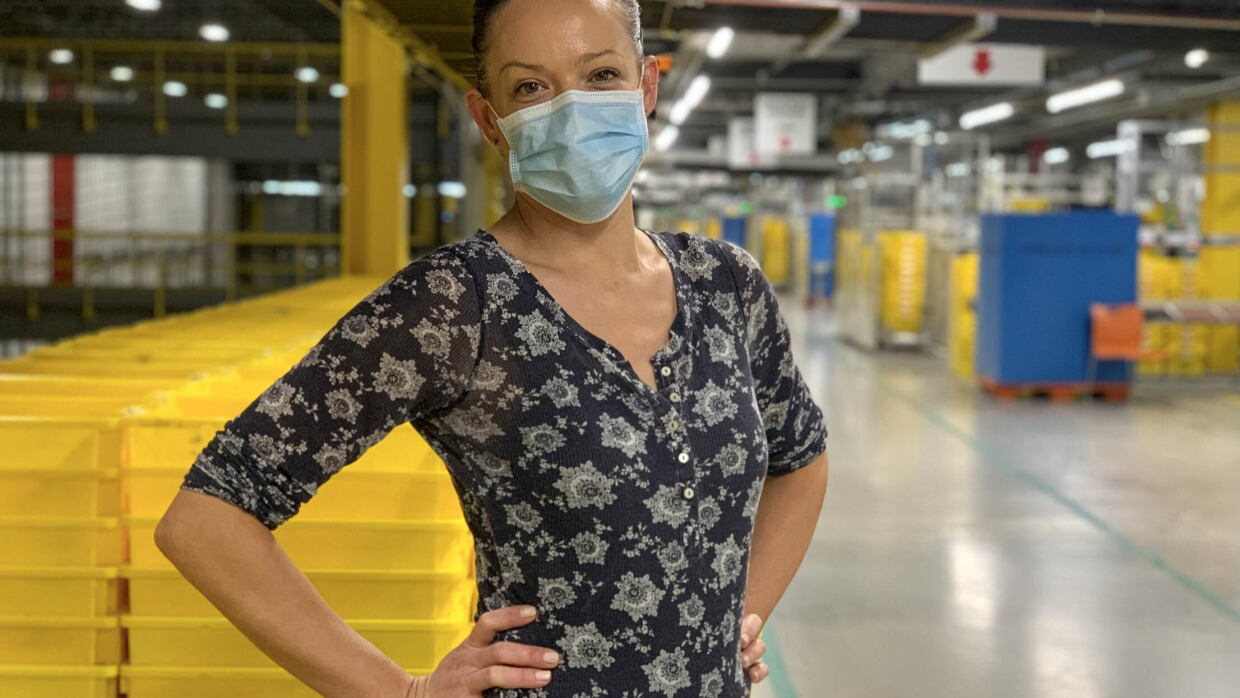 Una asociada está dentro del centro logístico. Lleva mascarila azu l, un jersey azul estampado con unoz dibujos de color blanco. Las mano las tiene en la cintura. De fondo cajas de color amarillo y azul.  