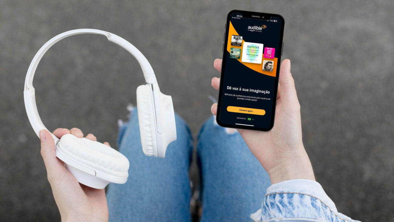 Um mulher segura um fone sem fio e um celular, com o aplicativo de Audible aberto
