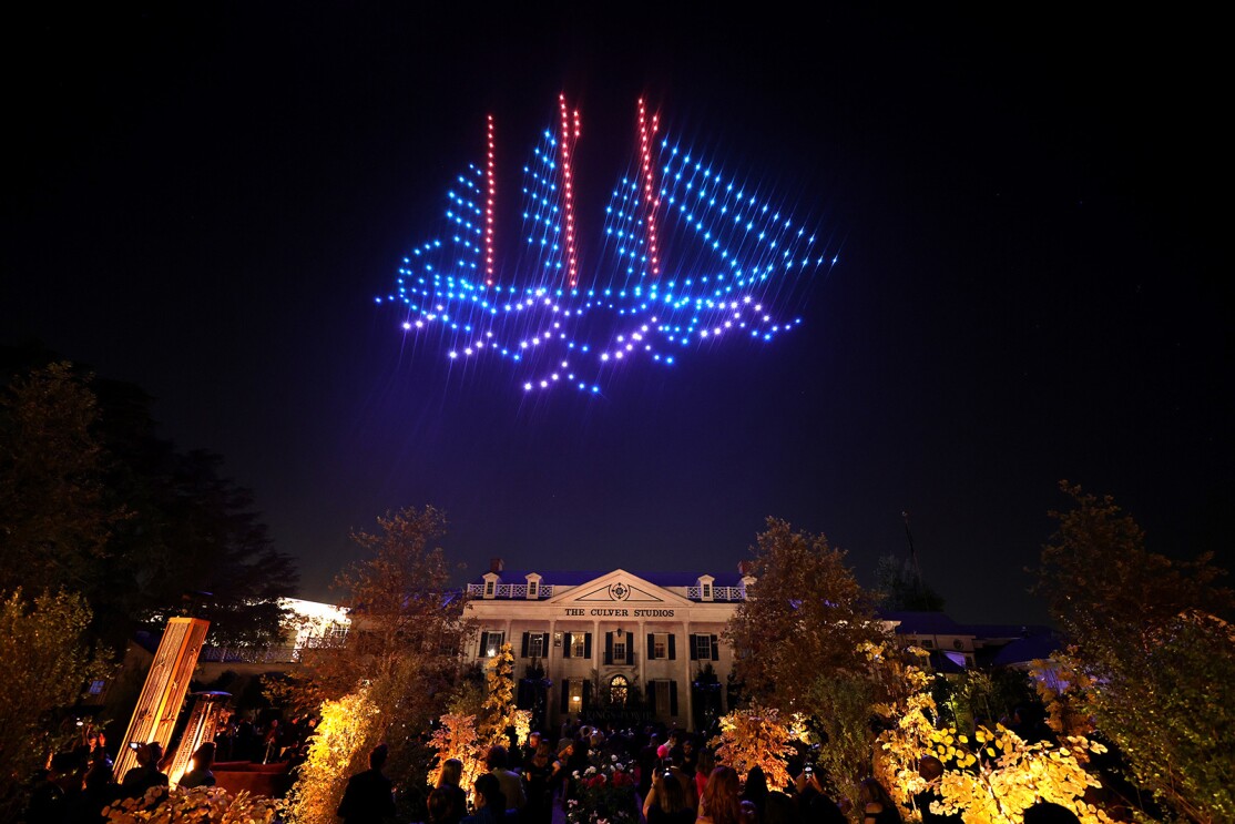 Red, blue, and purple drones light up the sky in the shape of a ship. On the ground below you see the Culver City Studios building and trees in the foreground.