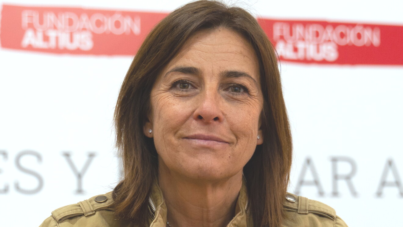 Natalia Rueda, directora de Comunicación de la Fundación Altius.  De fondo está un pared blanca con letras negras que no se puede apreciar ninguna palabra pero sobre fondo rojo está escrito en blanco Fundación Altius. Natalia lleva el pelo castaño por el hombro y suelto, tiene las manos cruzadas sobre la mesa. Lleva pulseras en la mano derecha, reloj en la izquierda y un anillo, las uñas de color rojo, dos collares, pendientes. Una chaqueta de color camel con dos bolsillo en la parte del pecho y debajo lleva un jersey negro con rayas blancas. 