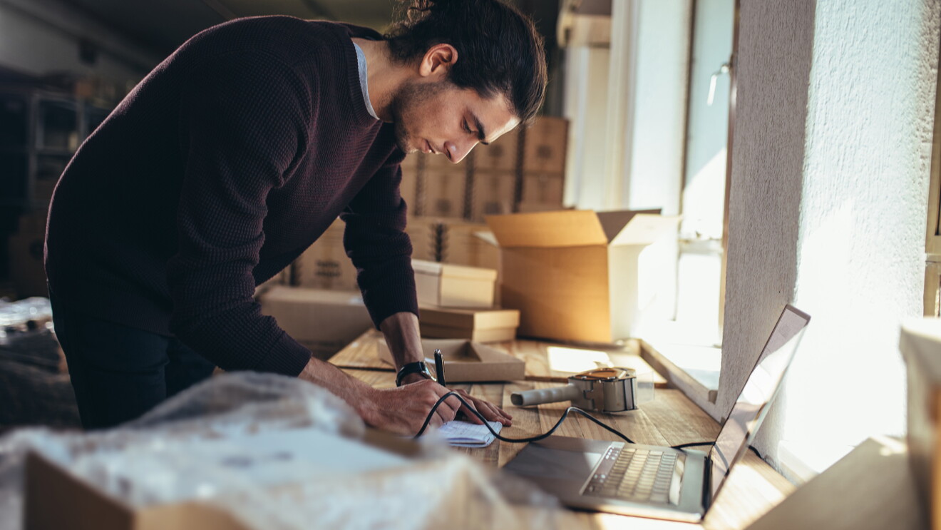 Man leaning over a desk, next to a laptop, with boxes in the background