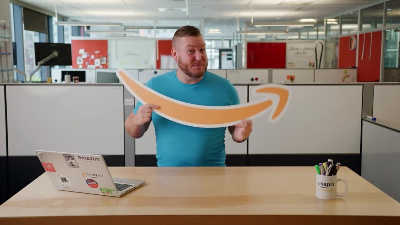 Responsable de selección de personal de Amazon DJ Cabeen agarrando la sonrisa-flecha de Amazon de cartón con las dos manos. El protagonista está delante de un mesa con un ordenador portátil y una taza de Amazon utilizada como lapicero. Lleva una camiseta de manga corta de color azul y tiene el pelo muy corto de los laterales y más largo arriba. De fondo mamparas divisorias de mesas. 