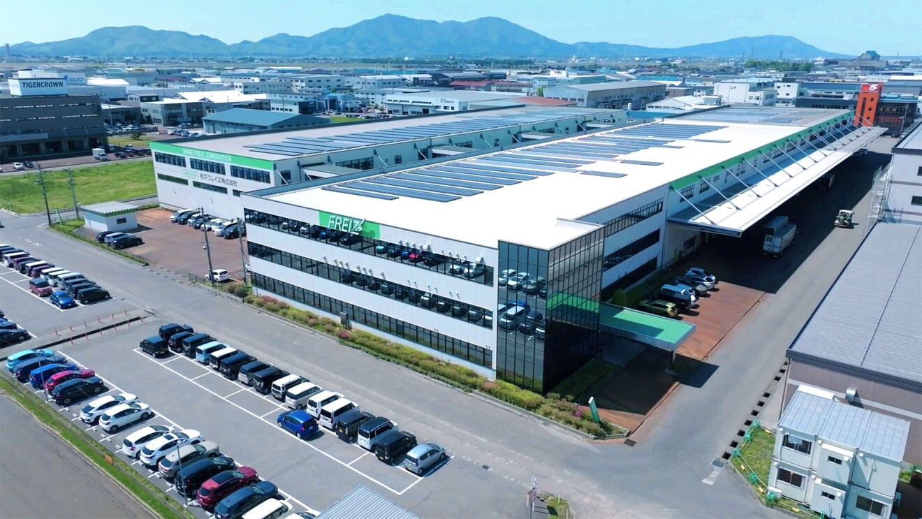3階建ての白い工場を斜め上から見下ろした構図。屋上には太陽光パネル、建物正面の壁に緑地に白で「FREIZ」のロゴ。手前の駐車場に多数の車。奥に青空と山並みが見える。