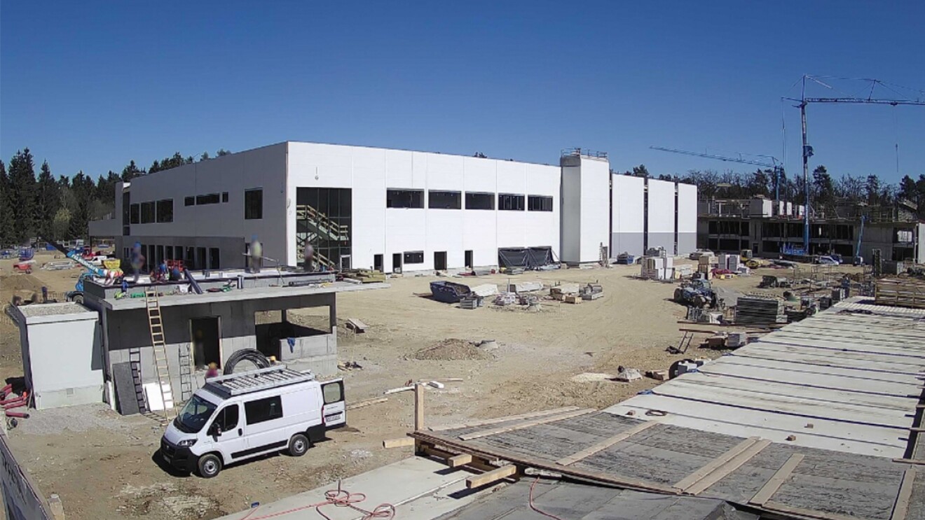 Baustatus des neuen Amazon-Verteilzentrums in Klagenfurt im März 2022. Ein weißer Van steht auf vor einem Gebäude, im Hintergrund ist das neue Verteilzentrum als weißes Gebäude zu sehen.