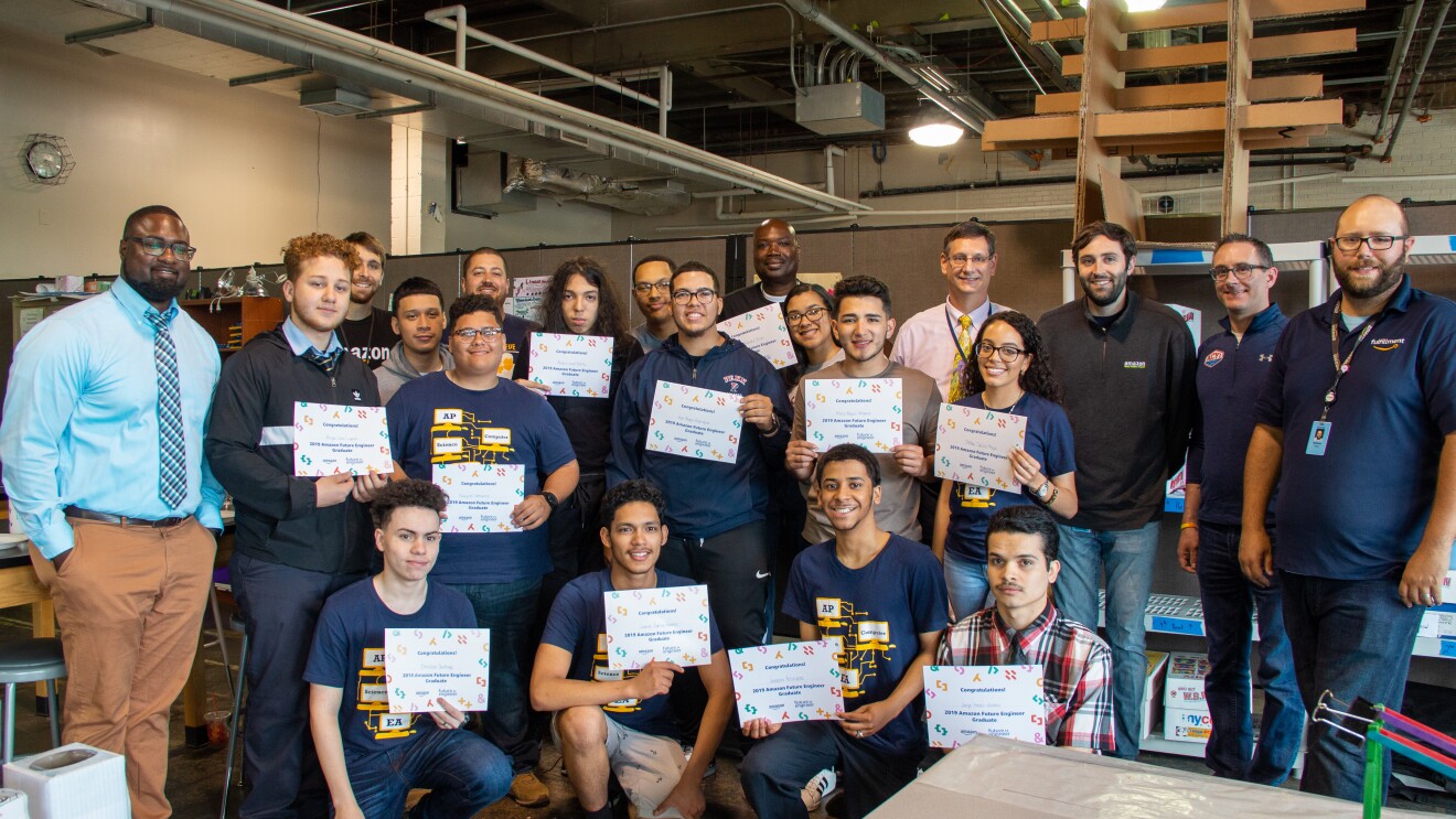 Several students gather together in a group at a high school. They hold up Amazon Future Engineer certificate, and are flanked by teachers.