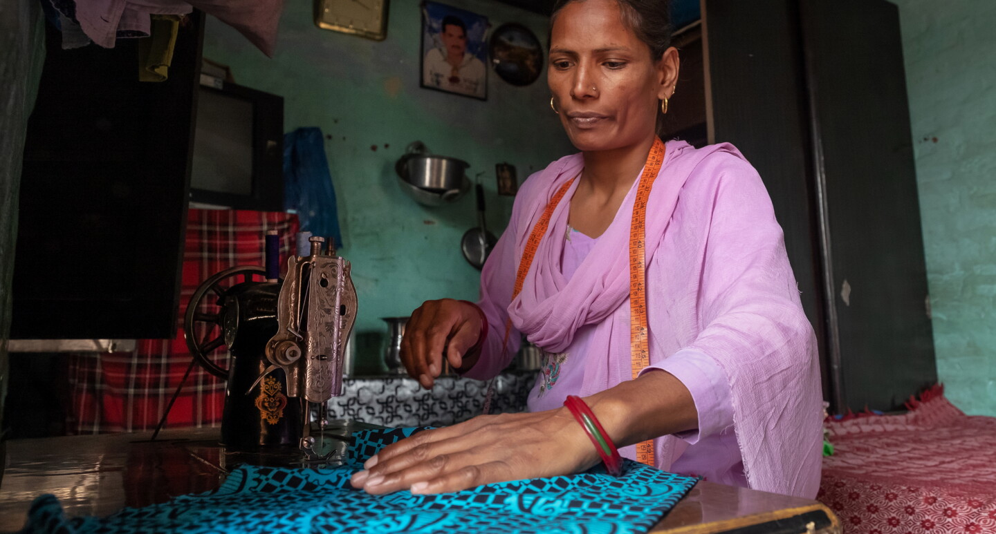 A woman sits at a table with a sewing machine. She has a measuring tape draped around her neck and uses her right hand to position a patterned fabric.