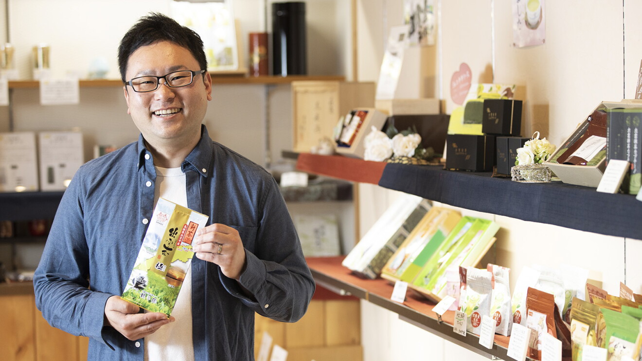 店内でお茶の袋を持つメガネをかけた男性が笑顔を見せている。ダンガリーシャツを着ている