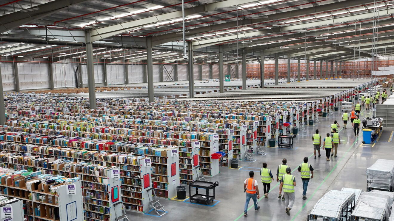 An image from the top of an Amazon fulfillment center. There are rows of shelves of products on the left of the photo and employees with their safety vests on walking alongside a walkway to the right of the photo.