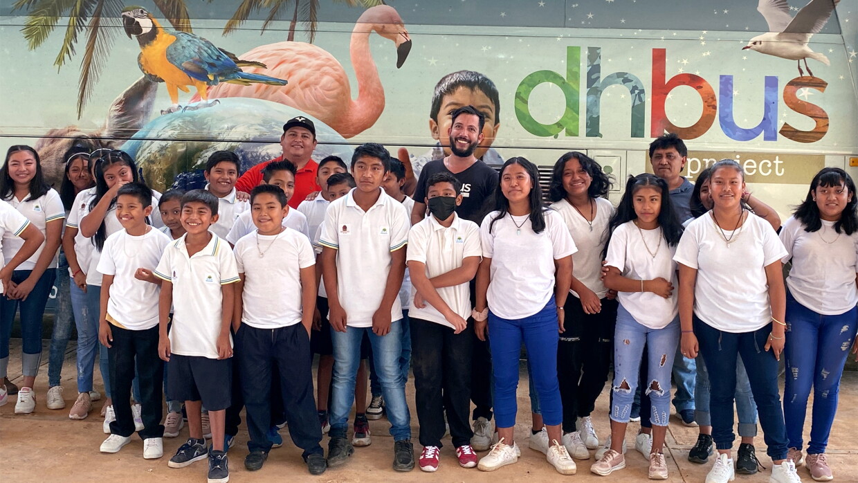 El DHBUS, un autobús acondicionado como aula itinerante, es un proyecto de innovación educativa diseñado para llevar programas a comunidades vulnerables y de difícil acceso. El bus está pintado con letras de colores dhbus project y pájaros exóticos típicos de México. Hay tres personas adultos que están en la parte trasera y delante, poco menos de 30 adolescentes vestidos con una camiseta blanca y jeans.  