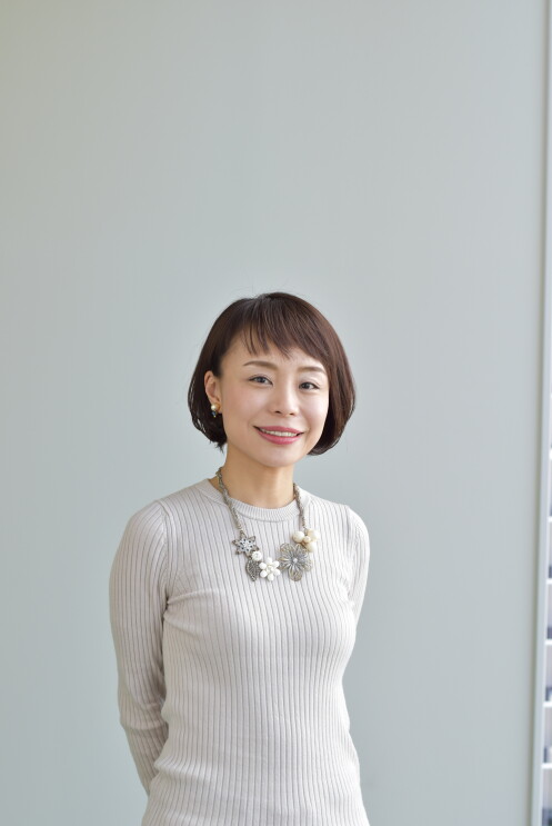 薄いグレーの壁の前に立ち、微笑む女性。白いトップスに、白とグレーの大ぶりな花のネックレス。