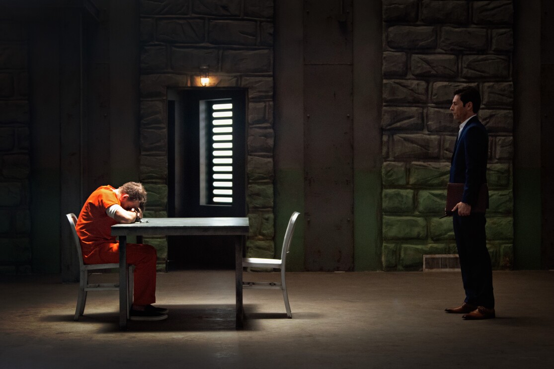 Prisonnier en combinaison orange affaissé à la table d'interrogatoire, homme en costume debout à proximité