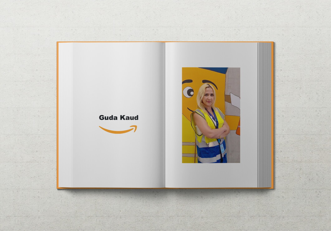 Guda, eine junge Frau mit blonden Haaren, lehnt in Sicherheitsweste an einer Wand auf dem das Amazon Maskottchen Peccy abgebildet ist.