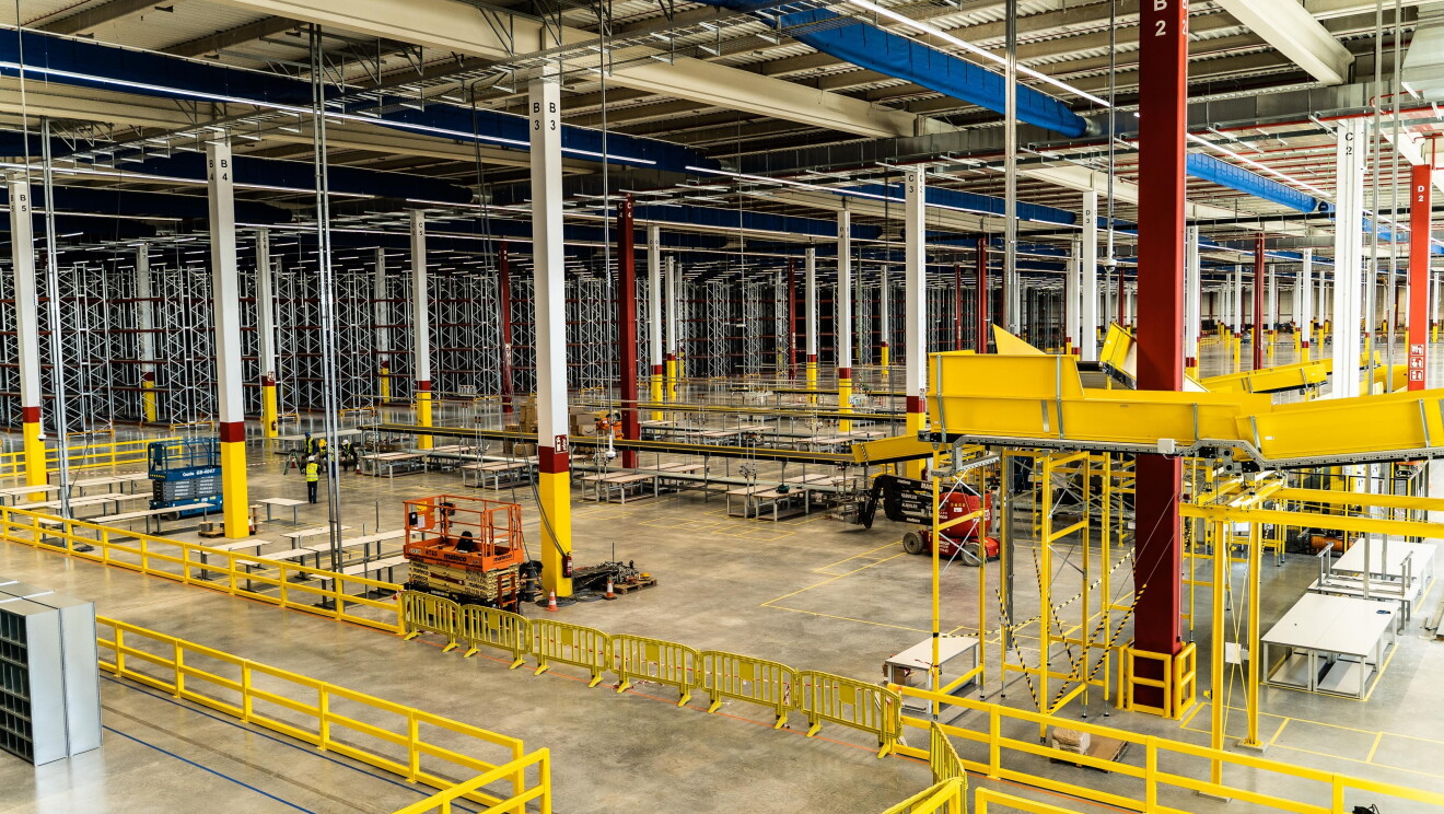Panorámica del centro logístico de Illescas. Se pueden observar grúas y trabajadores al fondo. En primer plano hay cintas transportadores y en el fondo estanterías para almacenar. 