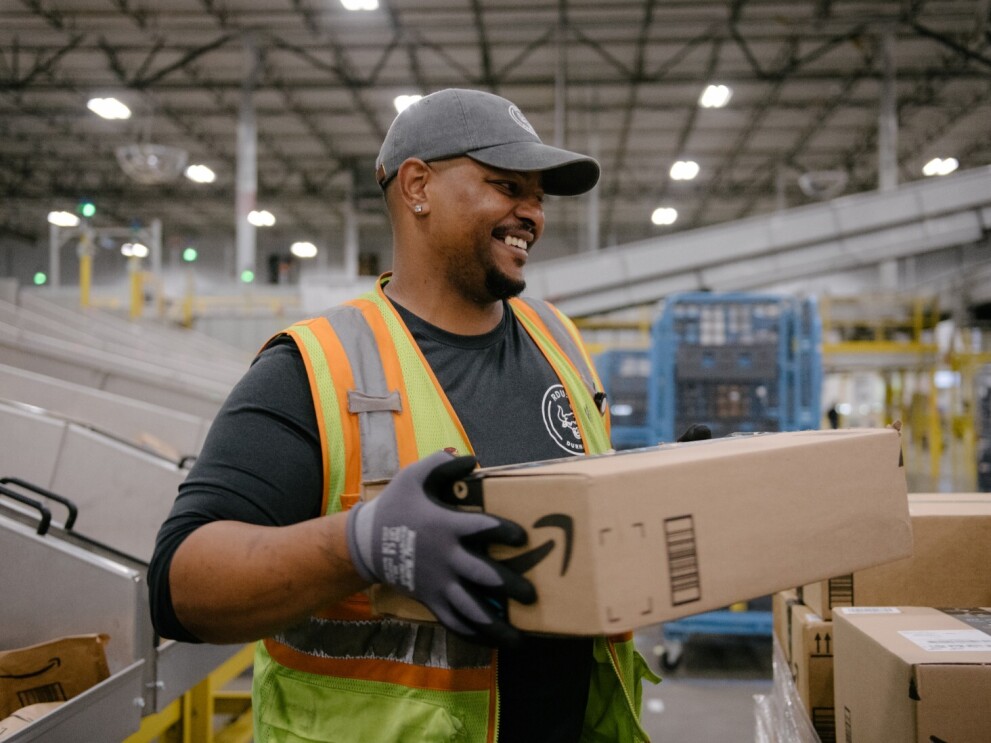 An Amazon fulfillment center employee wears a yellow safety vest and smiles as he moves an Amazon package.