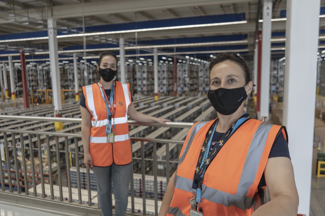 Marta Torrejón Serrano y María Jesús Serrano. Madre e hija posan en un balcón del centro logístico de Illescas que se ve de fondo. Están apoyadas a la banadilla manteniendo la distancia de seguridad. Las dos van con manga corta, mascarilla corporativa y chaleco naranja. Llevan el pelo recogido con una coleta.