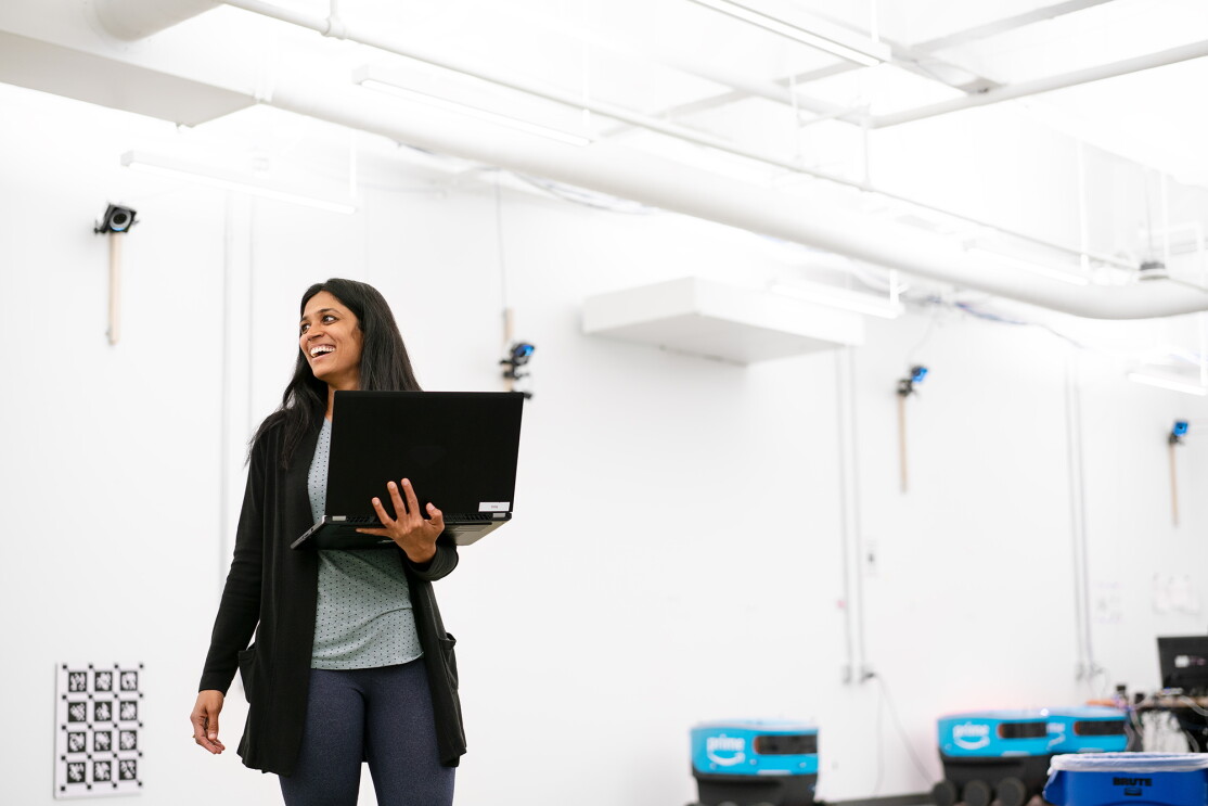 A woman stands in a white room, holding her laptop computer. Behind her, several Scout delivery robots are seen near the wall. 