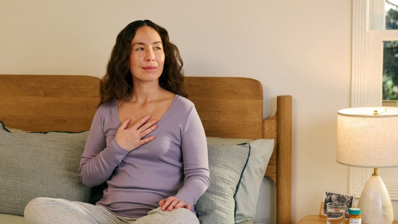 Woman in purple top resting hand on chest, sitting on bed