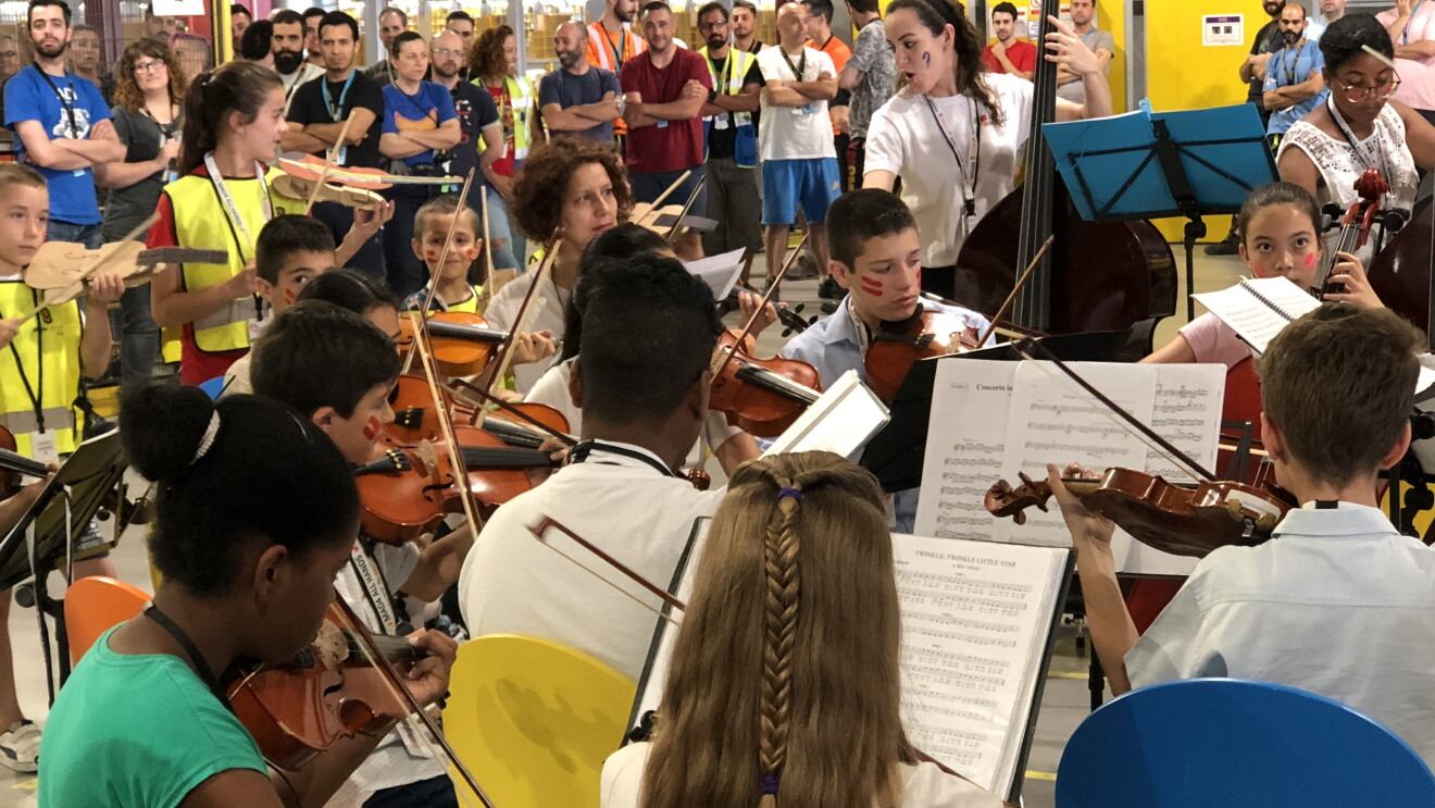 La camerata de Acción Social por la Música en un concierto en el centro logístico de San Fernando de Henares. En el fondo y de pie asociados disfrutando del concierto. En primer plano jóvenes sentados en sillas de colores y delante de la partitura tocando el violín y el violonchelo. También hay dos jóvenes tocando violines de cartón.