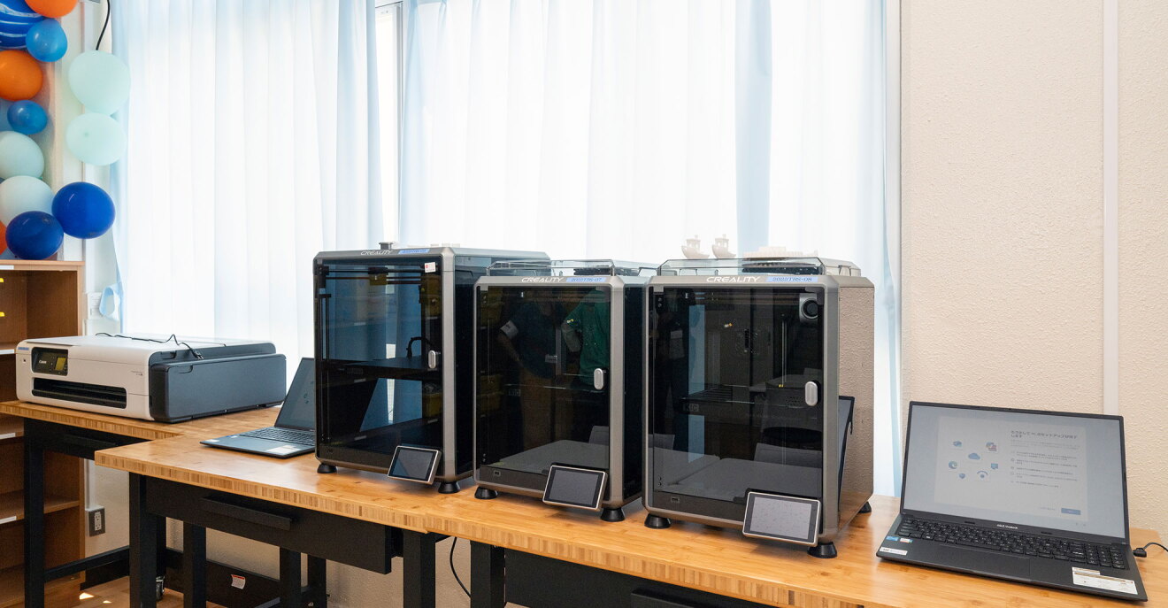 木製の作業台に3台の3Dプリンター、その両側にノートパソコンが1台ずつ、いちばん左側にプリンターが並んでいる。各3Dプリンターの前には小型のディスプレイパネル。壁際には白いカーテン。左上側にオレンジ、青、水色の風船で飾られている。
