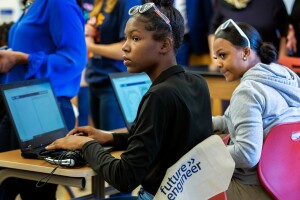 A photo of two students sitting at a desk in front of two laptops, listening to an instructor.