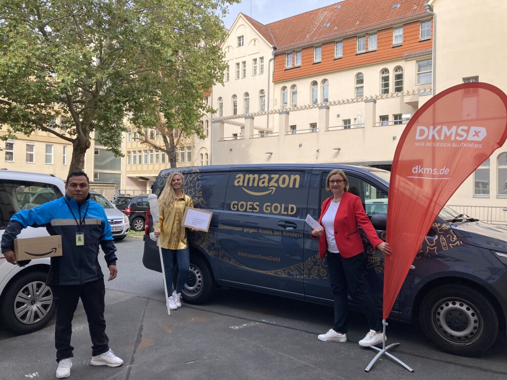 Amazon übergibt eine Spende an eine Vertreterin des DKMS. Die Vertreterin, eine Amazon Mitarbeiterin und ein Amazon Lieferpartner stehen vor einem mit "Amazon goes gold" beschrifteten blauen Liefervan.
