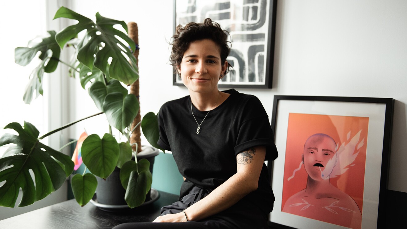 An image of Carmela Caldart smiling for a photo in a living room area. There is a plant and a framed illustrated image behind her.