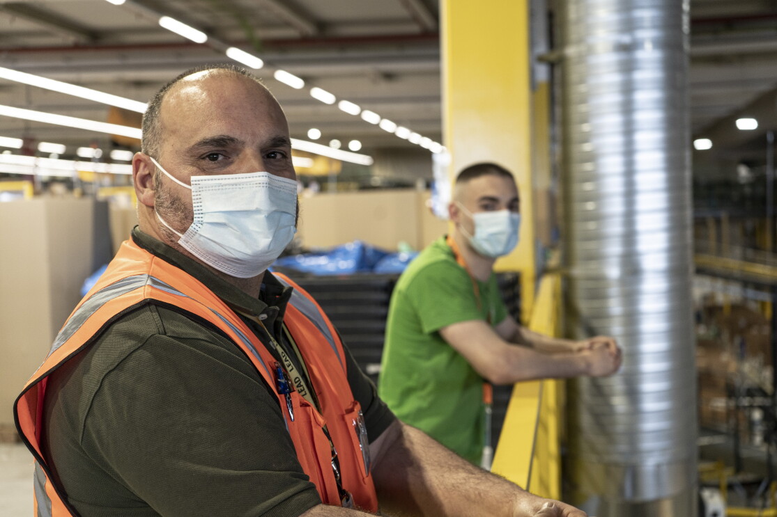Santiago And´res y Santi Andrés en el centro logístico de Amazon en San Ferando de Henares (Madrid). Desenfocado, de fondo, las estanterías amarillas de un centrlo logístico. En primer plano el padre, Santiago Andrés con pelo muy corto negro y blanco y un poco calvo. Lleva un polo de manga corta de color verde, chaleco naranja y mascarilla. Su hujo en segundo plano con camiseta verde más destacada y el badge con una cinta naranja y mascarilla. Los dos están  apoyados a la barandilla amarilla.