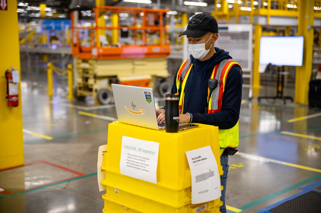 A man stands near a stack of yellow totes, typing on his computer in an amazon fulfillment center. 
