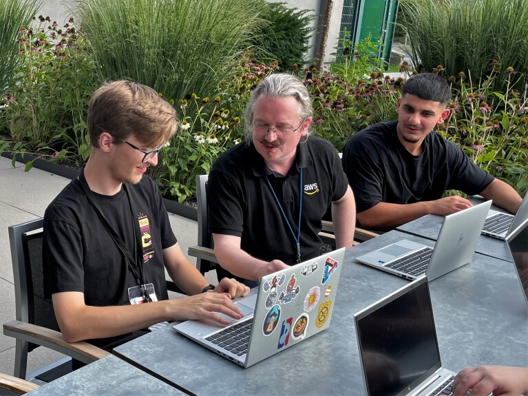Der AWS Ausbildungsleiter mit 2 Azubis an einem Terrassentisch im Freien. Alle haben Laptops vor sich.