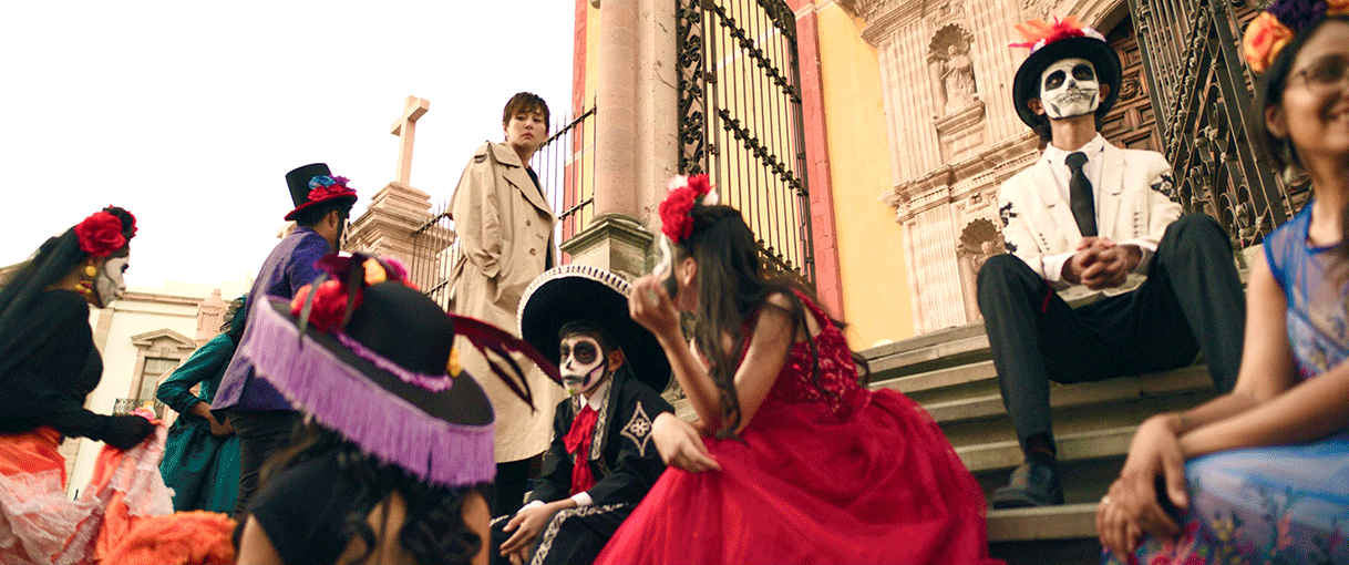 建物入口に向かう階段で、座ったり移動したりする仮装姿の人々。骸骨のフェイスペイントを施し、男性は黒や白のスーツに黒い帽子、女性はカラフルなドレスで頭に花飾り。