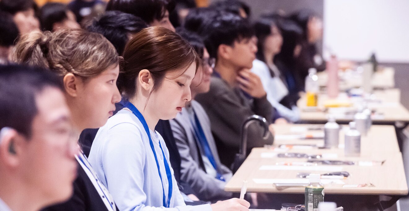 長机に並んで向かい、前を見つめる多数の若者たちの横顔。長机の上にはドリンクのボトルや筆記用具など。首に青いひもをかけている。