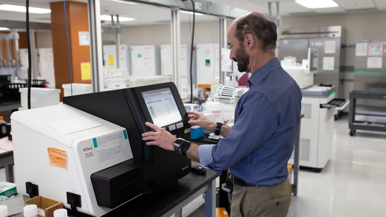 Michael Snyder, PhD, professor and chair of genetics for Stanford University’s School of Medicine stands in front of a machine with a monitor in his lab. He wears three different smartwatches on his wrists.