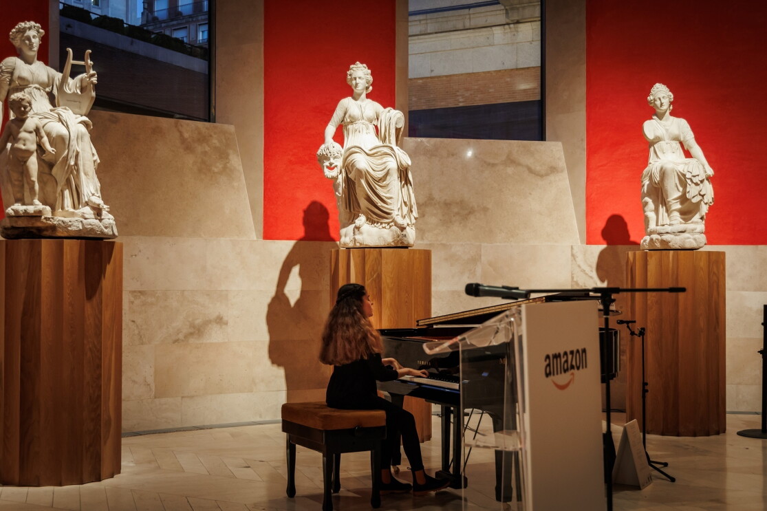 Piano Yamaha de cola negro con la tapa abierta. Plano general de Cristina Hernán Sanz, pianista residente del Museo del Prado tocando el piano en una sala ovalada del museo con tres escultura clásicas de mármol qu están colocadas en un pedestal de color madera. Detrás de cada escultura la pared es de color rojo. Hay dos grandes  ventanales. 
