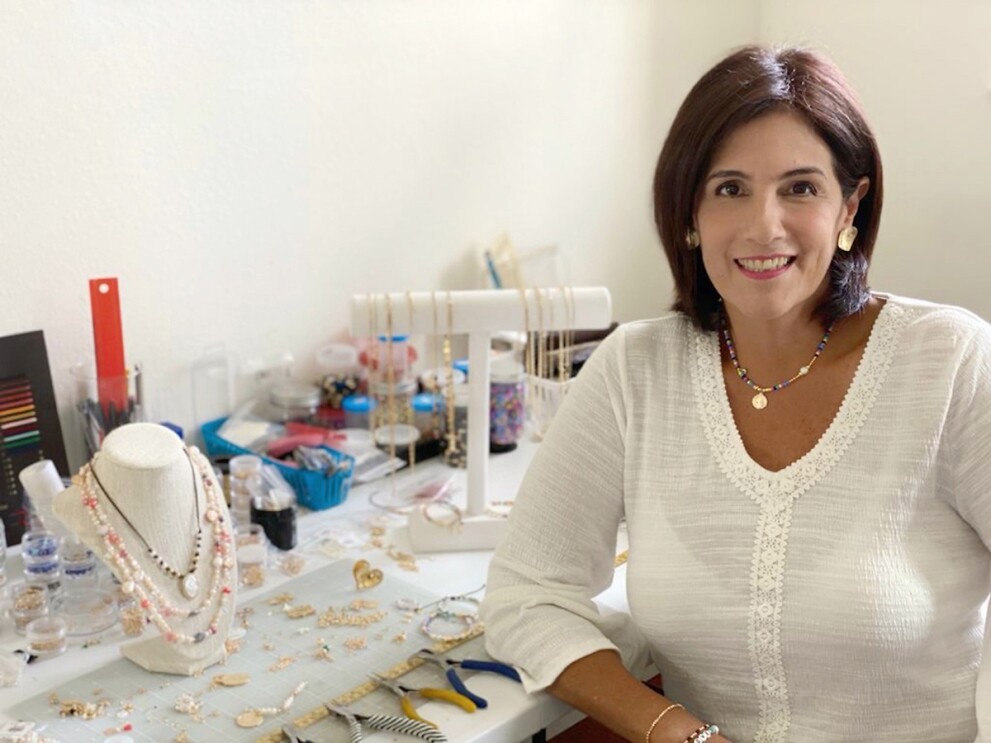 An image of a woman sitting in a white room with a jewelry-making stand in front of her. 