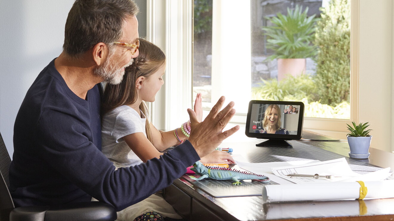 A grandfather sits in front of an Echo device with his granddaughter on his lap while they wave to someone they're calling on a video call.