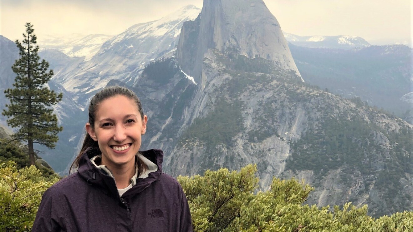 Caitlin smiles while standing outside. There is a mountain range in the near background.