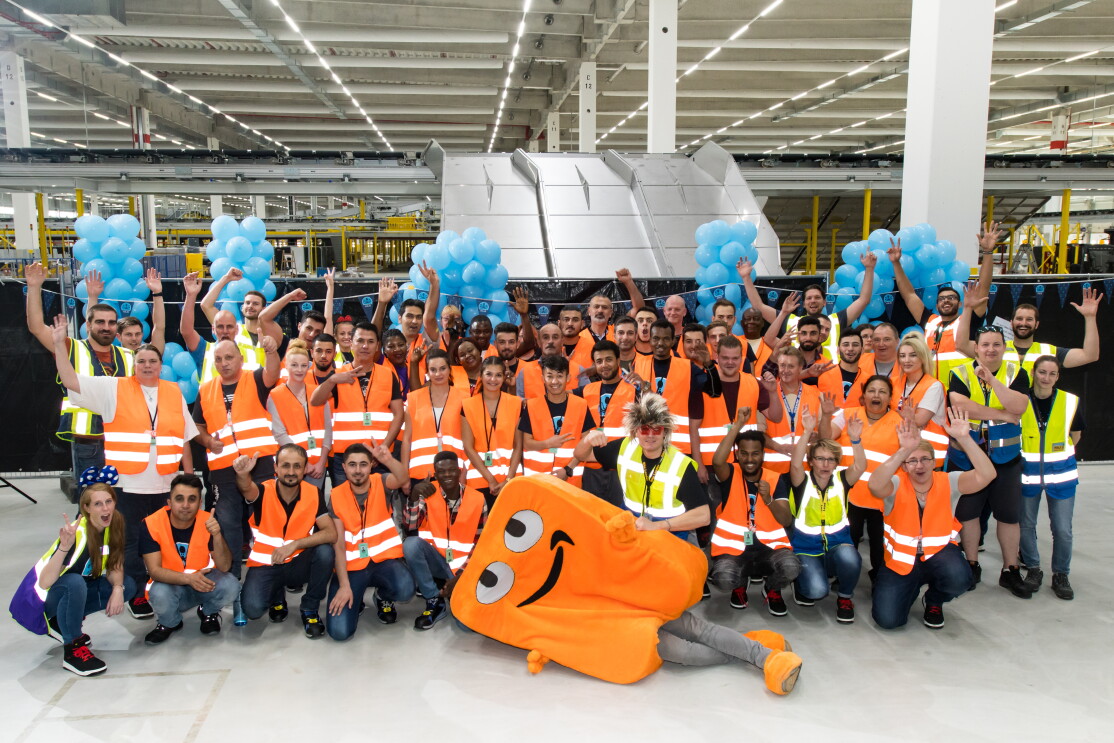 Gruppenbild der Mitarbeiter mit Maskottchen Peccy: Die Mitarbeiter tragen Sicherheitswesten, ein Mitarbeiter trägt das Peccy-Kostüm, das in orangener Farbe an einen Karton erinnert.