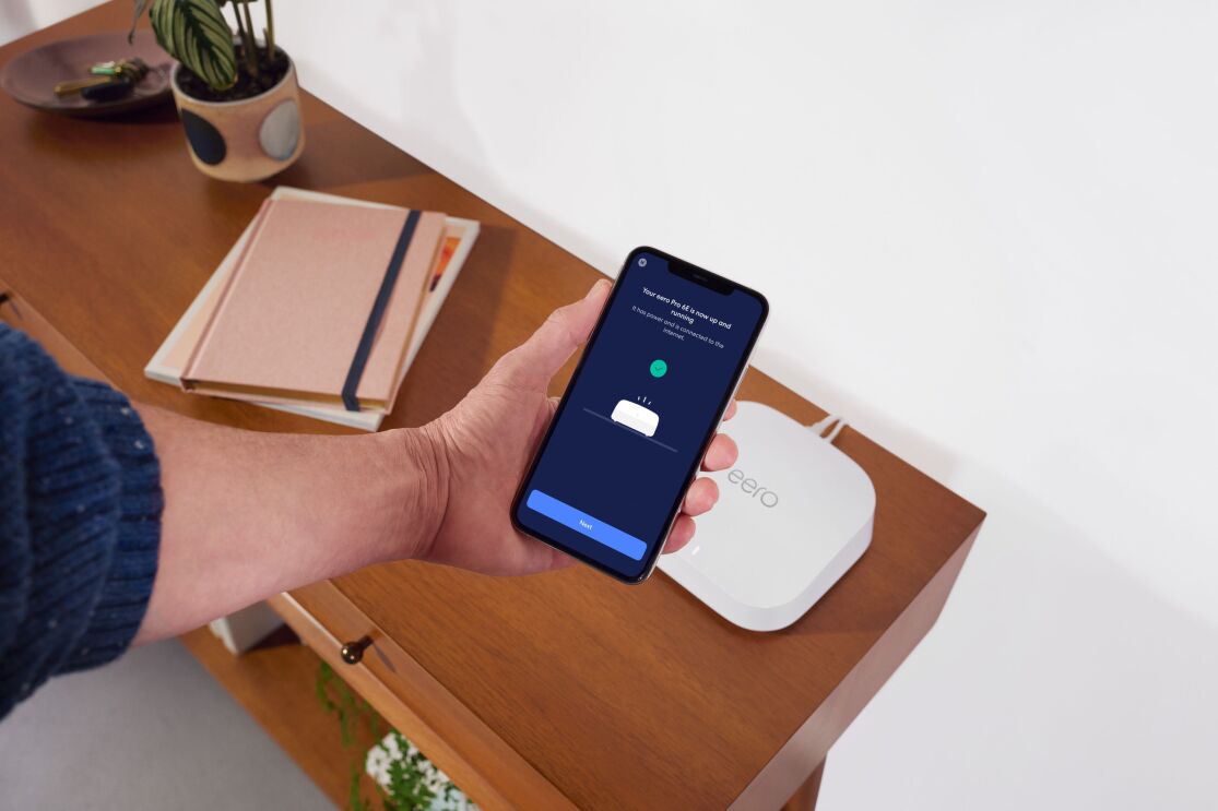 手で持った携帯の画面には白いデバイス(Eero)と繋がれたスクリーンが表示されている。そのデバイスは本や植物と一緒に机の上に置かれている。