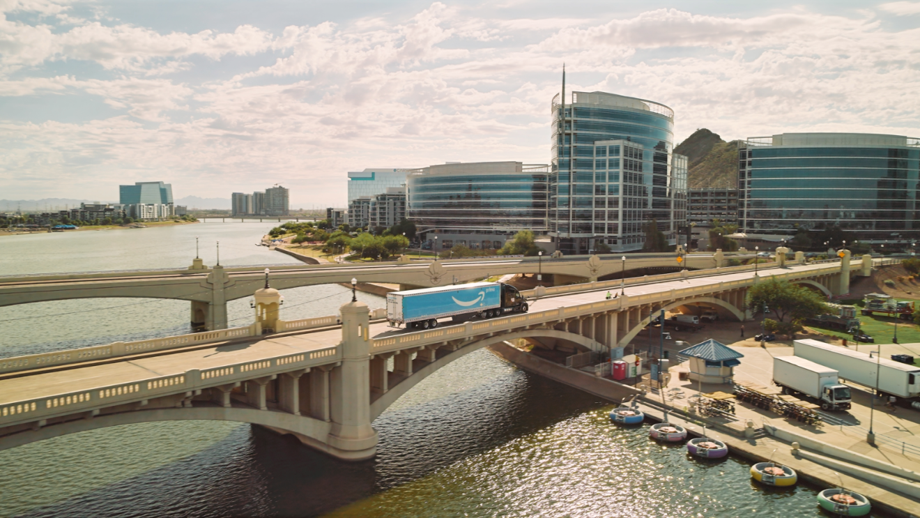 An Amazon Prime semi-truck crosses a bridge over a waterway with modern glass buildings in the background.