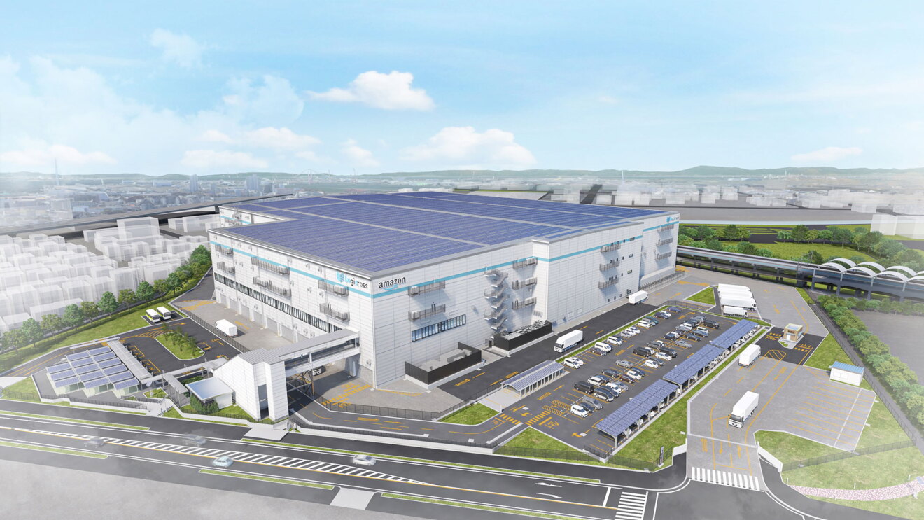 大規模物流施設の空撮イメージ。広大な屋根全面に太陽光パネルを配置。白い建物の側面に「amazon」の文字。周囲には駐車場、ソーラーカーポート、緑地帯、道路が整備され、建物前には大型トラック用の荷捌きエリア。背景には市街地と遠くに山々が広がる。
