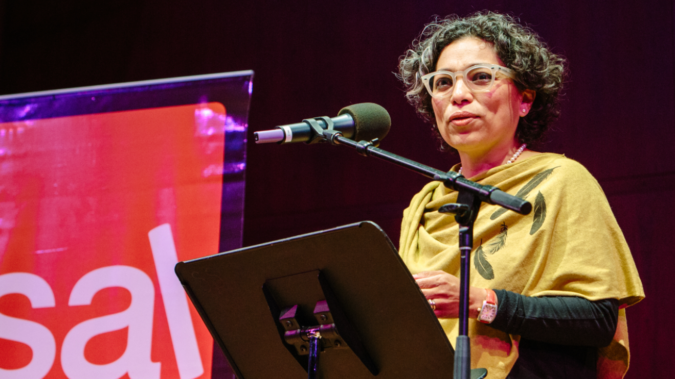 A woman standing at a lecture, in front of a portable podium and microphone. She appears to be speaking to a group, on a stage. In the background, Seattle Arts & Lectures logo is visible.