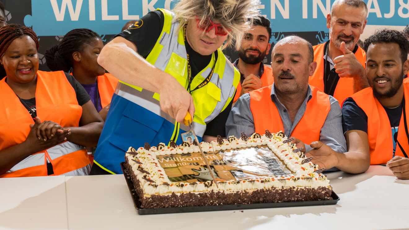 Ein Mann mit Igelperücke und roter Spaßbrille, von Logistikmitarbeitern umgeben, schneidet eine rechteckige Torte an