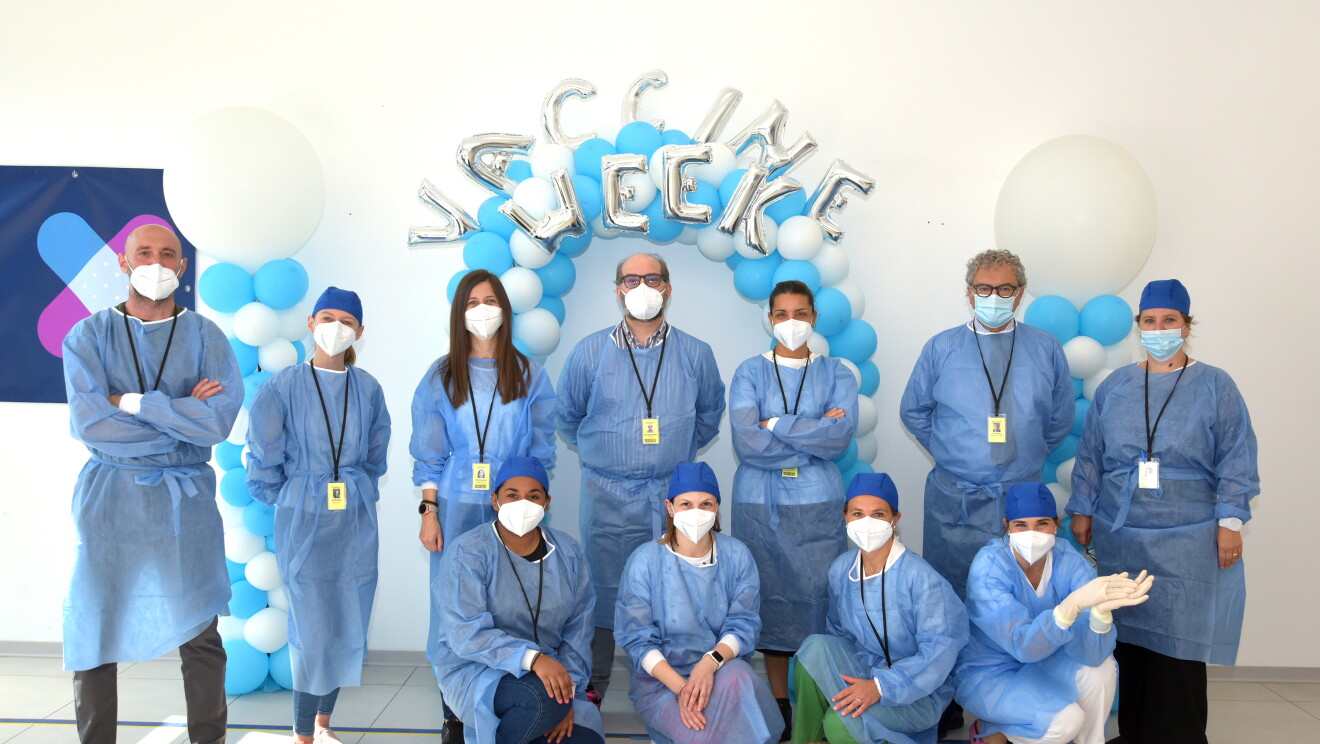 Un gruppo di medici in tute protettive blu posa davanti a due archi di palloncini bianchi e blu con la scritta 'Vaccine Week' alle loro spalle