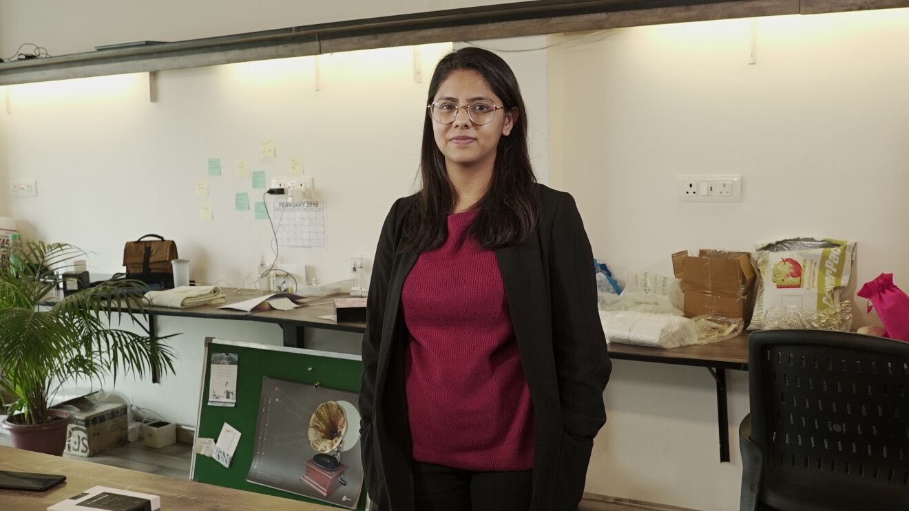 Tanvi Johri, creator of the all natural sanittary napkin, Carmesi, stands in her office wearing a tee-shirt and trousers topped with a jacket. Behind her is a long table with packages and huge potted plant stands to her right