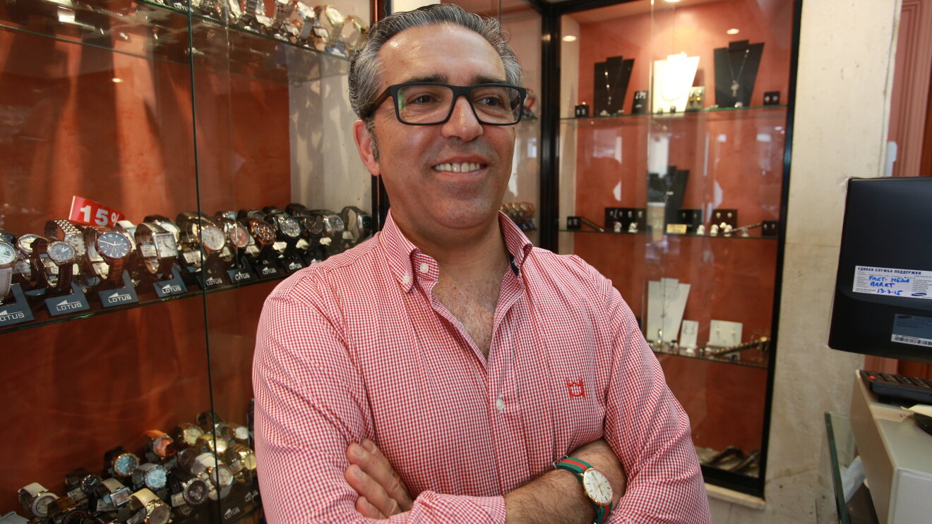 José Antonio Marín, de Marín Joyeros, es un hombre de mediana edad. Con una camisa roja y blanca y gafas de pasta, posa en el mostrador de su tienda delante de un escaparate de relojes. 