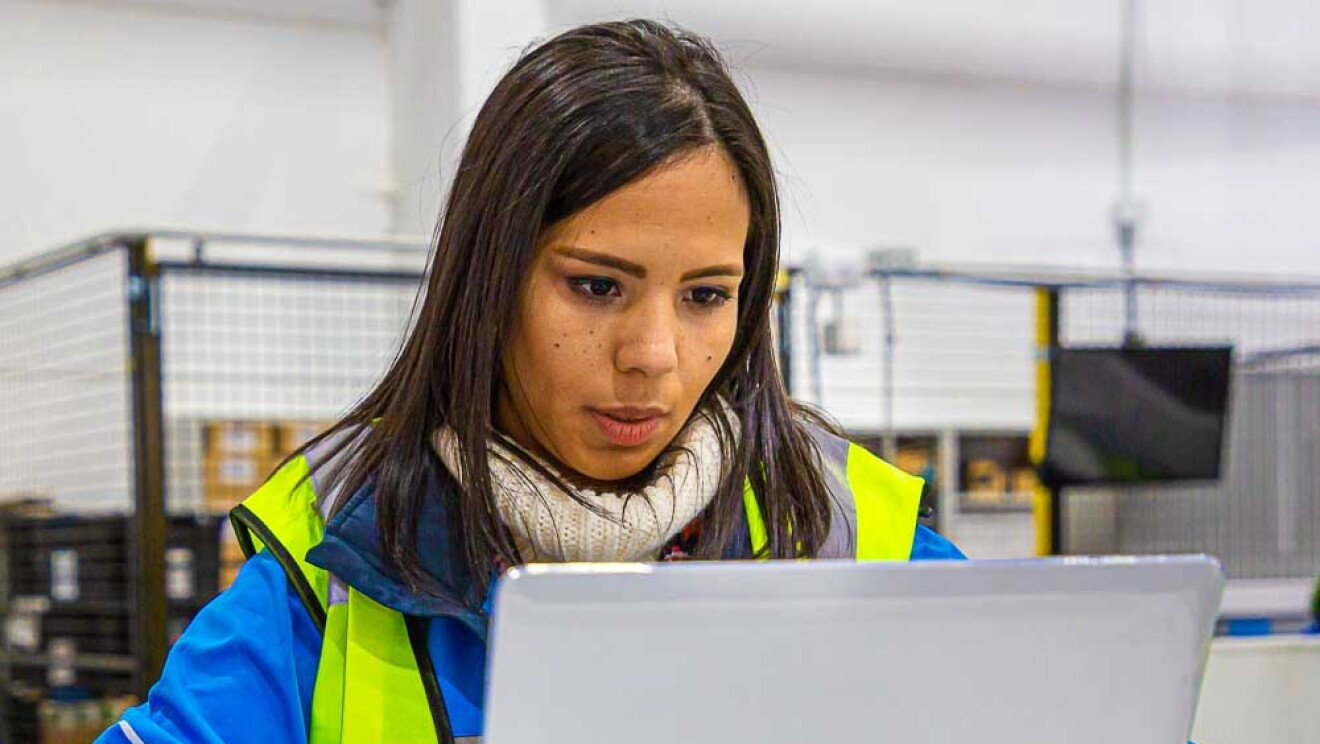 Ivette Duran, CEO de Canaima Logistics, trabajando en un centro logístico de Amazon. Está sentada en una mesa trabajando con un ordenador. Tiene el pelo largo de color negro y es morena de piel. Lleva un jersey de color blanco y encima una chaqueta azul con el logo de Prime Now y un chaleco fluorescente. 