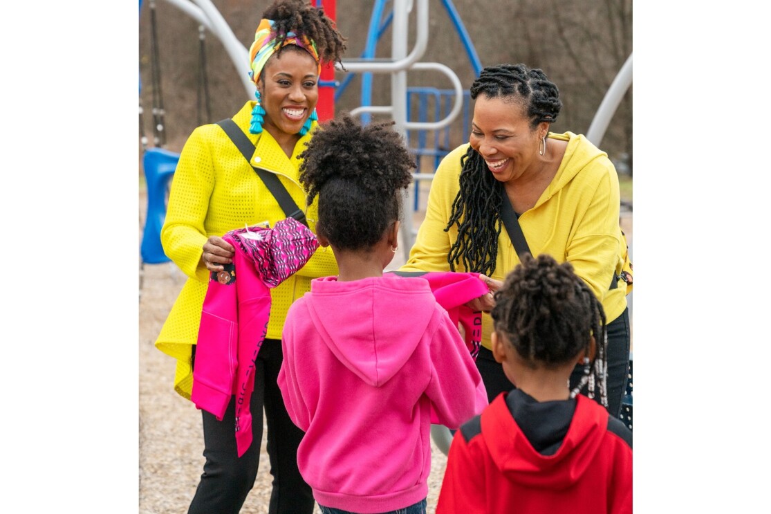 Cara and Jenae smile as they give children at a playground some of their apparel to wear.