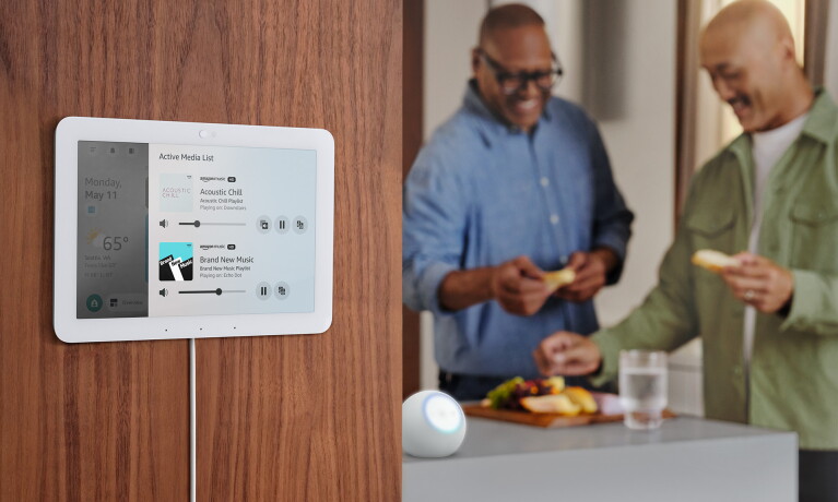 Amazon Echo smart home display mounted on wooden wall showing active media controls and weather widget while two people prepare food in kitchen