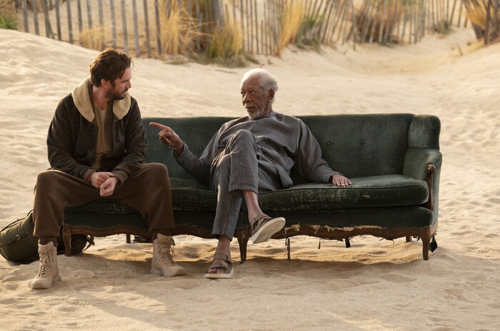 An image of Morgan Freeman and Dan Stevens sitting on a couch on a beach talking to each other. Freeman is pointing at Stevens casually as he speaks. 