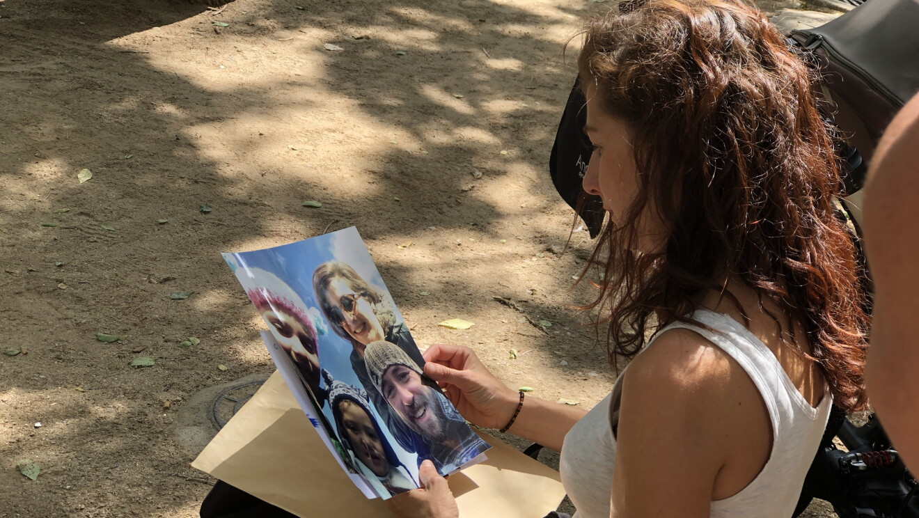 Irene sentada en un banco de un parque observa una fotografía de su familia. La sujeta con las dos manos. Viste con un jersei blanco sin mangas y una falda negra larga hasta los pies. 